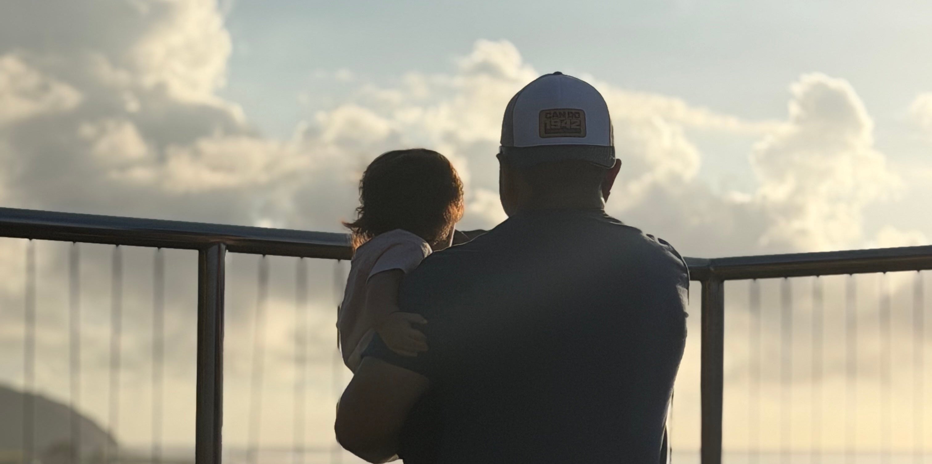 Person holding a child by a railing with a scenic view of water and clouds.